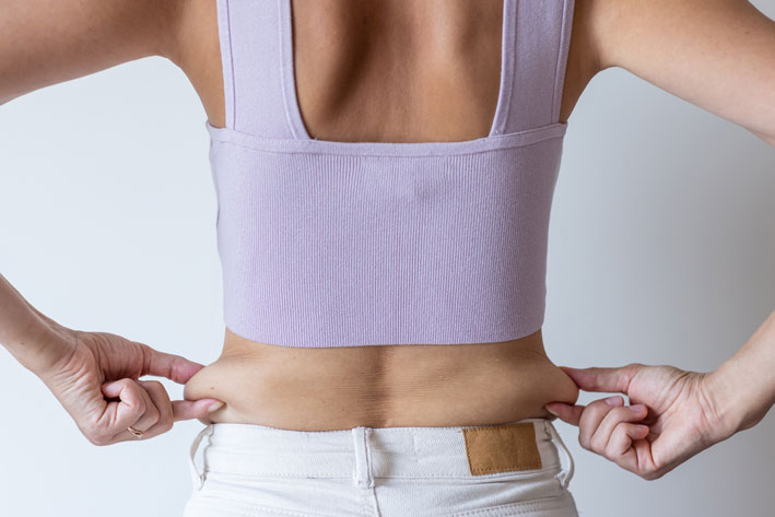 Massive Weight Loss | Services 2 Cropped image of overweight woman, tucking, hiding fat naked big excessive belly with navel in white jeans. Dangling down stomach, big size tummy. Drag away of abdomen. Go on diet, liposuction.